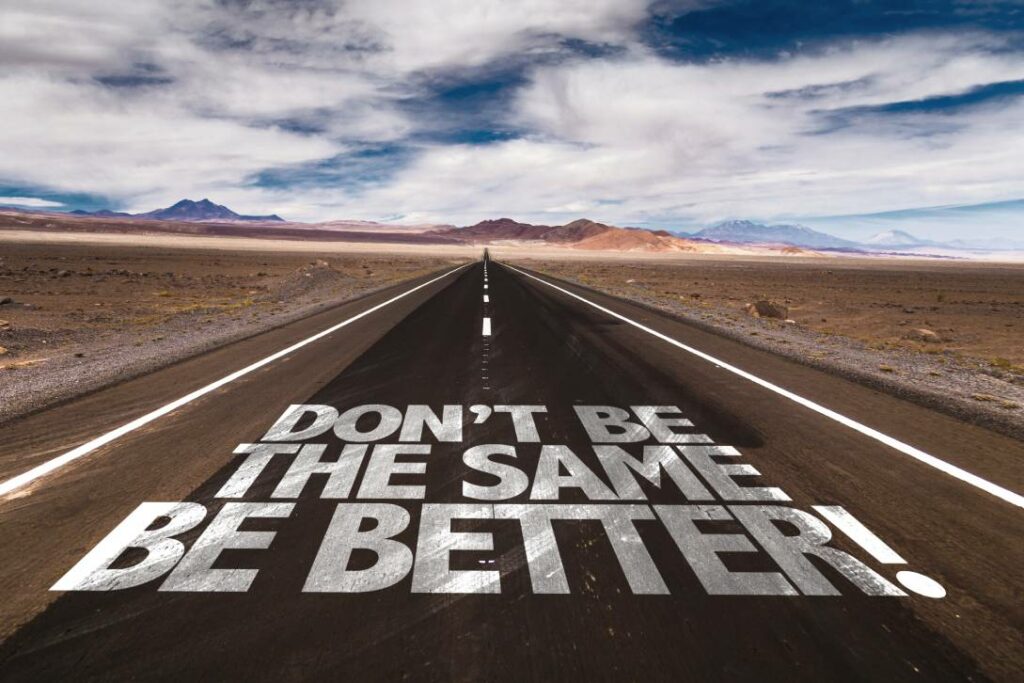 En rak ökenväg med "DON'T BE SAMMA, BETTER!" målat med stora vita bokstäver på asfalten, berg och moln i bakgrunden.
